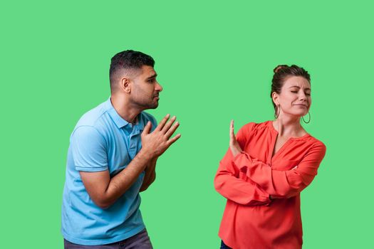 Portrait of funny couple in casual wear standing together, man holding arms in prayer and pleading forgiveness, looking with hope at naughty woman, isolated on green background, indoor studio shot