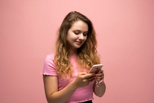 Smiling young woman girl in pink t-shirt using mobile phone and typing sms message to a friends isolated on pink wall background studio. People lifestyle concept. Mock up copy space