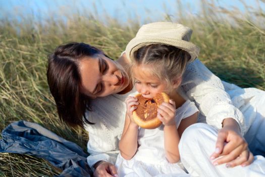 Mother and daughter sitting picnic high yellow grass child eat bun. Happy family with one child resting nature outdoor mom with little girl 5 years old. Countryside