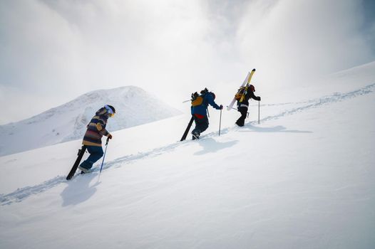 Three friends snowboarders skiers go uphill with a snowboard and skis in their hands for backcountry or freeride against the backdrop of the snow-capped mountains of the Alpine resort. Ski tour concept with group of peoples