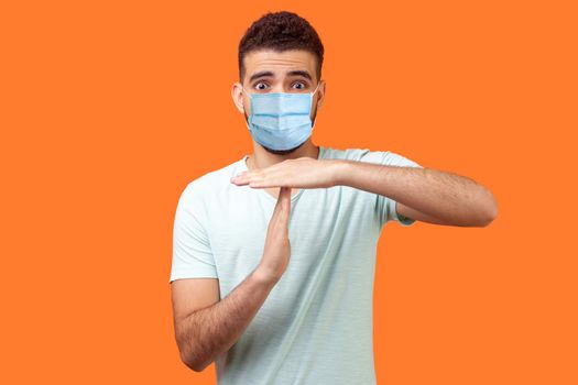 I need more time. Portrait of frustrated brunette man with surgical medical mask showing time out gesture, looking with imploring eyes, hurry to meet deadline. studio shot on orange background