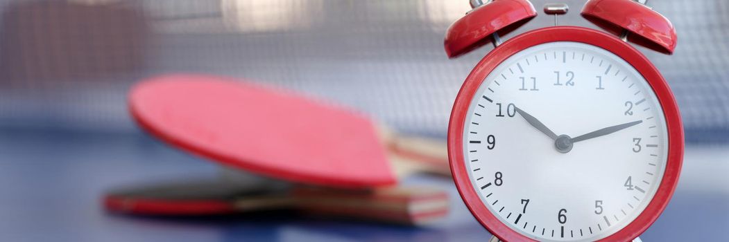 Red alarm clock on a tennis table, close-up, blurry. Office ping pong during the break. Sports recreation for employees