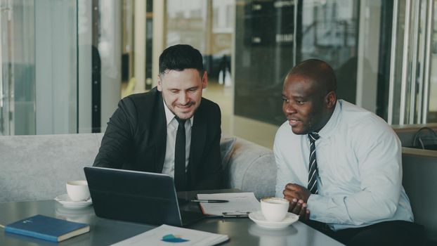 Two multi ethnic businessmen looking at laptop computer and laughing in glassy cafe. Men having fun and joking. African American businessman and Caucasian man feel success.