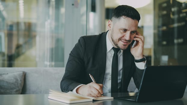 Portrait of cheerful Caucasian businessman sitting, smiling, talking on smartphone with his friend and writing down information in notepad while looking at laptop in modern cafe during lunch time