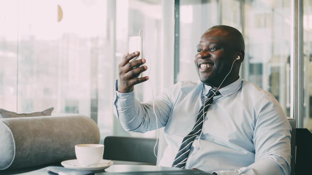 African American businessman in formal clothes smiling, laughing and talking to family through smartphone camera in cafe. He waves his hand and gesticulates happily with earbuds in ears