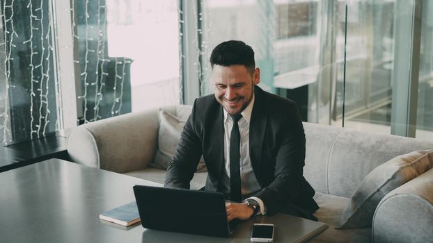 Caucasian businessman in formal wear smiling while surfing the net on his laptop in classy cafe during lunch time