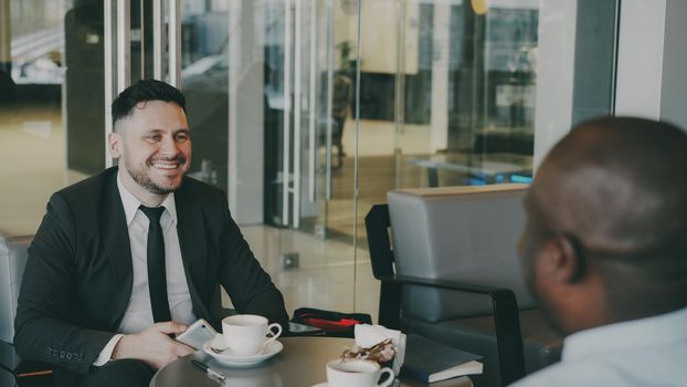 Two multi-ethnic businessmen sitting, smiling and discussing their startup in airy cafe during lunch time. Cheery African American partner strokes his knees happily. Coffee cups are on their table