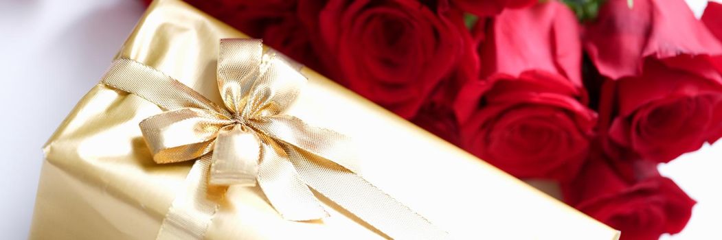 Gift wrapped in gold and a bouquet of red roses, close-up. Beautiful fighting box with ribbon, floristry