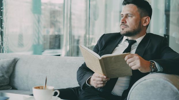 Successful Smart Caucasian businessman in formal clothes reading text book with and drinking coffee in classy cafe at lunch break