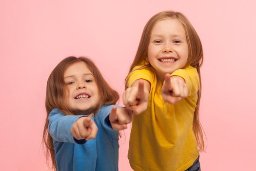 Hey you. Two charming happy little girls pointing to camera, indicating lucky one and smiling, choosing goods in child store, we need this concept. indoor studio shot isolated on pink background