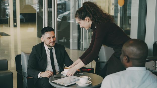Optimistic bearded Caucasian businessman in formal clothes smiling and discussing business profit with his African American colleague in cafe at lunch time. Cheery waitress bringing cup of coffee.
