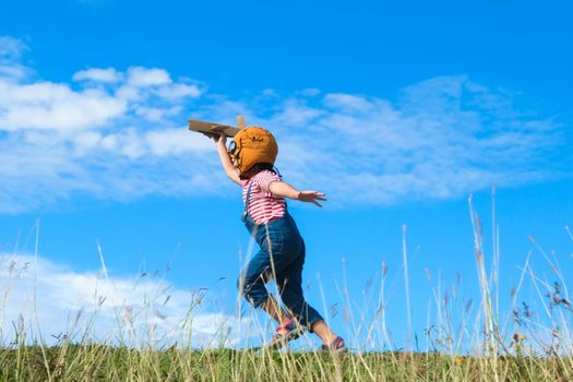 Cute little girl running through the meadow on a sunny day with a toy plane in hand. Happy kid playing with cardboard plane against blue summer sky background. Childhood dream imagination concept.
