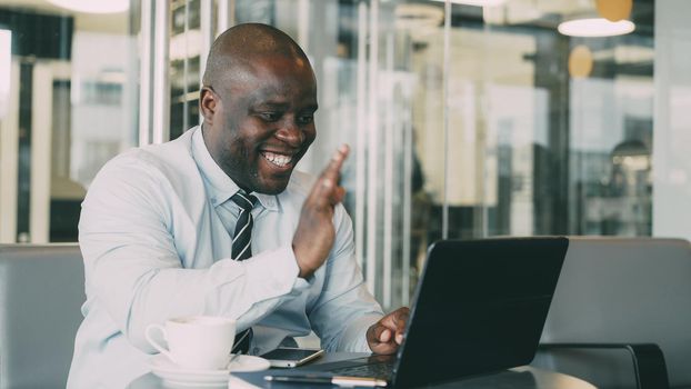 African Americanbusinessman in formal wear having online video call with his laptop and talking to his family in classy cafe during lunch break. He smiles and waving his hand cheerfully.