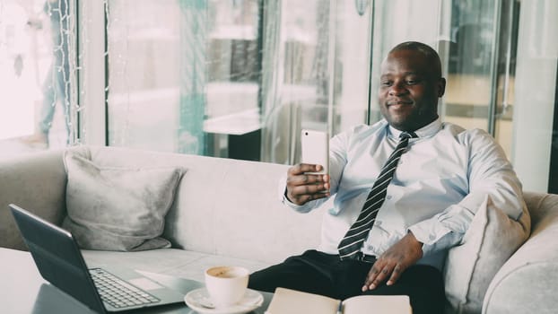 Portrait of cheerful African American businessman taking a selfie on his smartphone and showing a thumb up gesture while drinking coffee in modern cafe. He smiles happily and optimistically