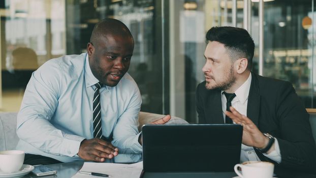 Two business colleagues looking at laptop computer and discussing their startup plan in modern office with glass walls. Bearded businessman and his partner sitting at table and talking future deals