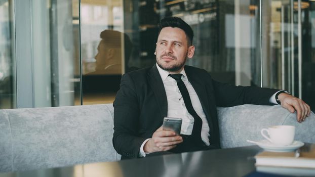 Smiling bearded Caucasian businessman in formal clothes smiling, relaxing and looking at his smartphone screen while drinking coffee in airy cafe during lunch time