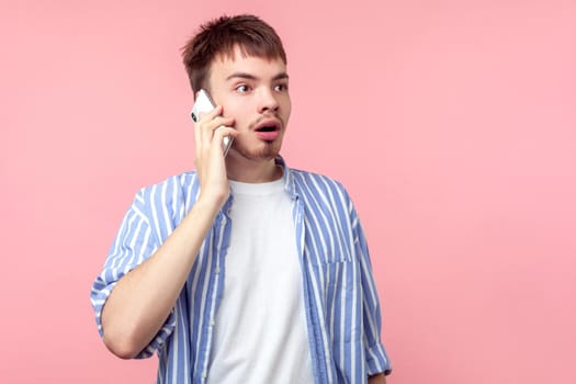 Portrait of astonished young brown-haired man with small beard and mustache in casual shirt talking on phone, looking aside with shocked face expression. indoor studio shot isolated on pink background