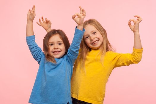 Children showing okay, everything is fine. Two charming happy little girls gesturing ok with fingers and smiling together, meaning all right, we agree. indoor studio shot isolated on pink background