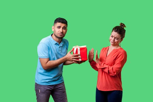 Portrait of upset resentful man in casual clothes giving present to naughty beautiful woman, dissatisfied girl showing refusal gesture, bad gift. isolated on green background, indoor studio shot