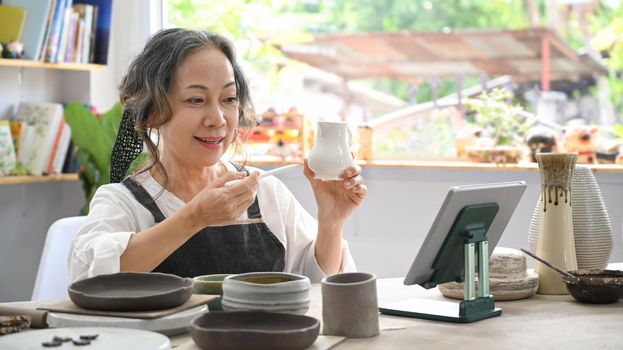 Concentrated middle aged woman painting pottery bowl in workshop. Indoors lifestyle activity, handicraft, hobbies concept.