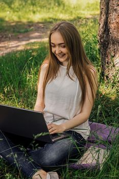 On the banner, a young girl works with a laptop in the fresh air in the park, sitting on the lawn. The concept of remote work. Work as a freelancer. The girl takes courses on a laptop and smiles