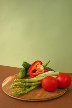 Vegetables lie on a wooden board: tomatoes, asparagus, cucumbers, red bell peppers. brown, light green background. place for text