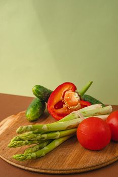 Vegetables lie on a wooden board: tomatoes, asparagus, cucumbers, red bell peppers. brown, light green background. place for text