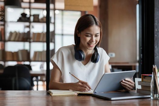 Asian female student with digital tablet, studying remotely listening to an online lesson, taking notes in a notebook, while sitting in a coffee shop. Online education.