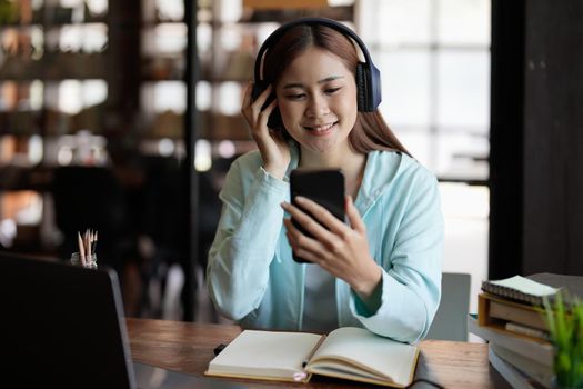 Happy asian student girl in wireless headphones using learning app on mobile phones, watching webinar on smartphone, listening to audio course, studying at open book, copy book at home.