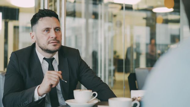 Bearded Caucasian entrepreneur gesticulating and discussing his startup plan with investor in formal wear in glassy cafe during during meeting