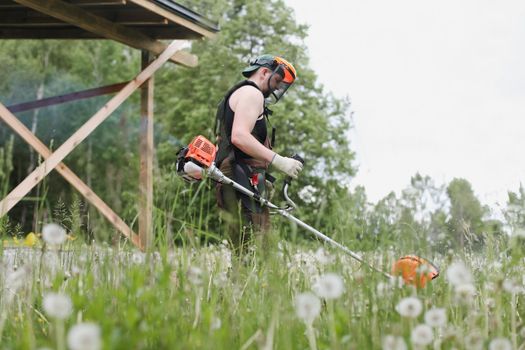 Worker man mowing tall grass with petrol lawn trimmer in the garden or backyard. Process of lawn trimming