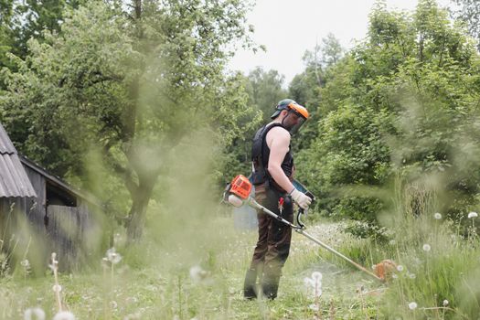 Worker man mowing tall grass with petrol lawn trimmer in the garden or backyard. Process of lawn trimming