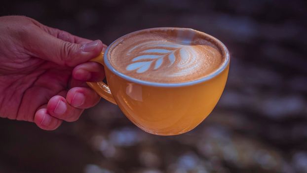 Hand holding a yellow cup of latte coffee top view showing latte art in leaf shape