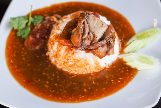 Red barbecued pork on rice, food serve with sweet curry and vegetable (cucumber, coriander) on white ceramic plate.