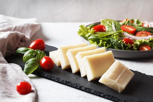 A piece of fresh sliced halloumi on a slate board with cherry tomatoes and spinach leaves. Preparing to cook cheese on the grill.