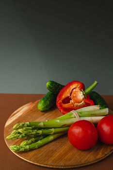 Vegetables lie on a wooden board: tomatoes, asparagus, cucumbers, red bell peppers. brown, dark gray background. place for text