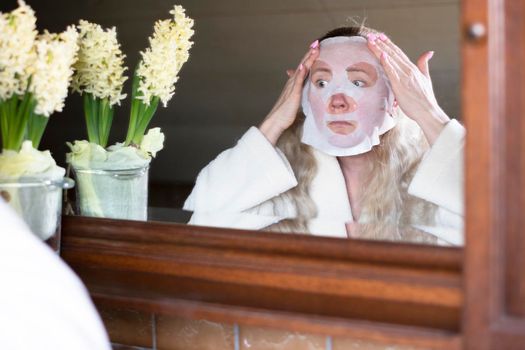 a middle-aged woman is busy with morning hygiene, trying in vain to straighten the mask with cleansing and moisturizing impregnation on her face High quality photo