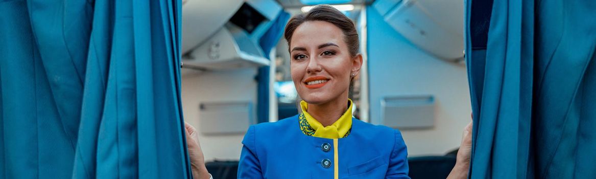 Joyful flight attendant in air hostess uniform looking at camera and smiling while holding drapes in airplane cabin