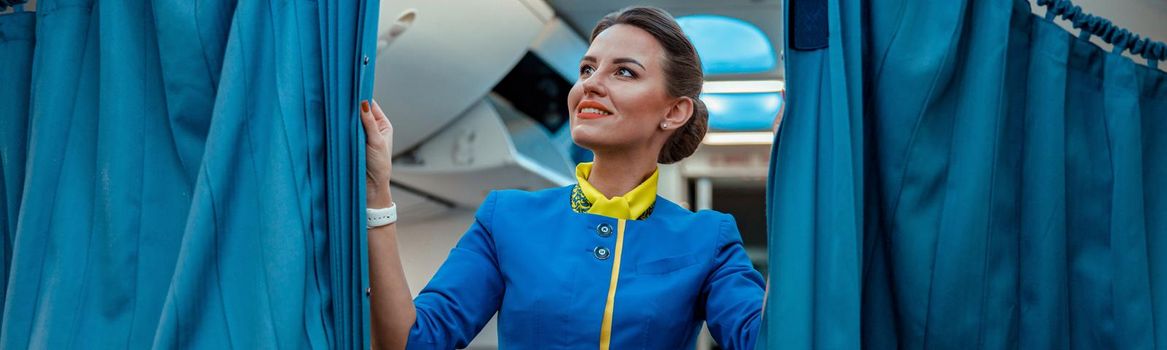 Cheerful flight attendant in air hostess uniform looking at drapes and smiling while standing in airplane cabin