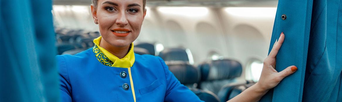Cheerful flight attendant in air hostess uniform looking at camera and smiling while holding curtains in airplane