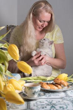 blonde middle-aged hugging a kitten and drinking tea, still life with yellow tulips and cakes a stack of books on the table. High quality photo