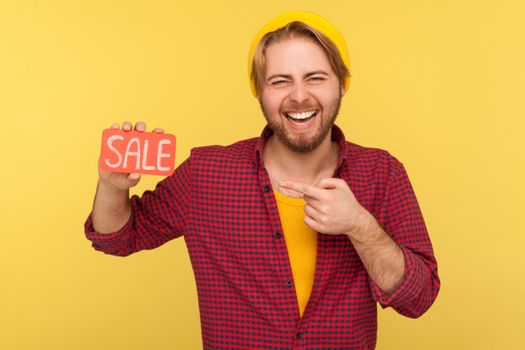 Excited trendy hipster guy in checkered shirt pointing at Sale word and laughing loudly, feeling carefree amused by discounts, Black Friday low prices. indoor studio shot isolated on yellow background