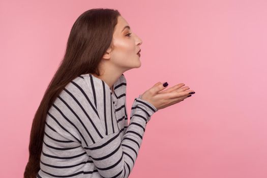 Love you. Side view of sensual attractive woman in striped sweatshirt sending air kiss over palms, expressing fondness, romantic feelings and flirt. indoor studio shot isolated on pink background