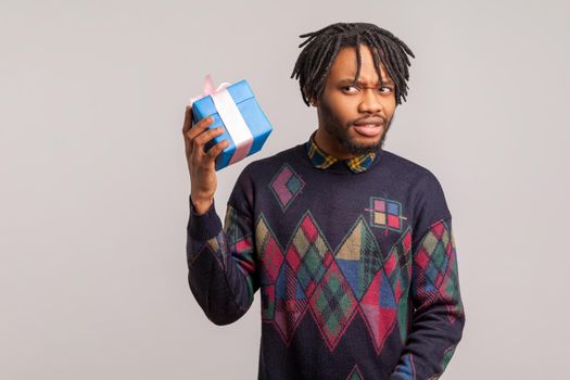 Disappointed curious african guy with dreadlocks trying to guess what is inside gift box, sudden present, delivery service. Indoor studio shot isolated on gray background