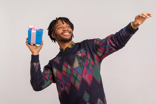 Positive african man with dreadlocks in casual style sweater holding present box and smiling going to throw it, concept of express delivery service. Indoor studio shot isolated on gray background