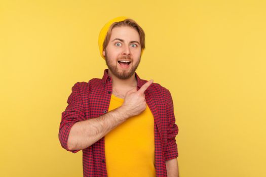 Look, advertise here. Surprised excited hipster guy in beanie hat looking at camera with happy amazed expression, pointing away at copy space, blank yellow background for commercial idea. isolated