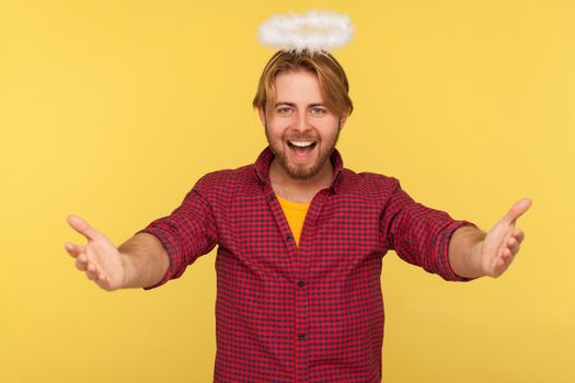 Come into my arms. Funny amiable bearded guy with saint nimbus over head stretching hands to camera and smiling friendly, going to embrace, share love. indoor studio shot isolated on yellow background