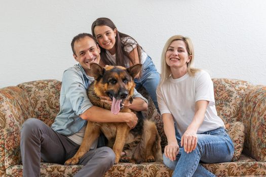 Beautiful young parents, their cute little daughter looking at camera and smiling, sitting with their cute dog on sofa at home.