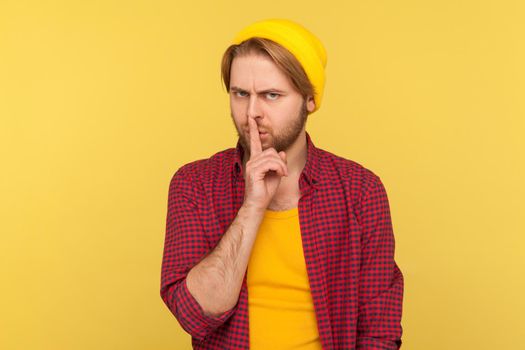 Shh, be quiet. Hipster bearded guy in beanie hat and checkered shirt shushing with finger on lips hand gesture, asking for silence secrecy, mute turn down sound. indoor studio shot isolated on yellow
