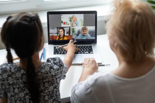 Portrait of happy grandmother and little granddaughter making video conference on pc sitting at table, waving hands at screen, greeting somebody, chatting with parents, enjoying online communication